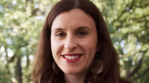 A woman with long dark hair and big earrings smiles at the camera.