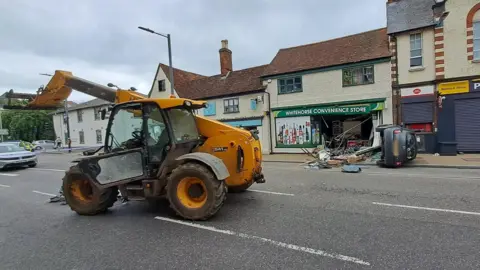 Contributed A yellow JCB telehandler sits in the middle of a street outside a damaged shop front. A grey car is on its side in front of the shop. There are a row of buildings and a police car to the left, parked behind police tape. 