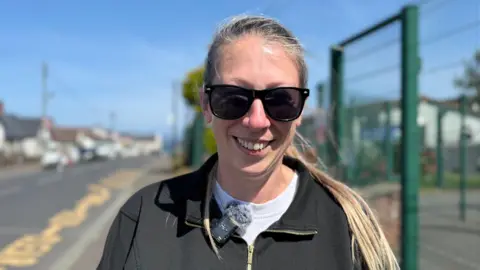 The image shows a woman standing outdoors on a bright, sunny day, smiling warmly at the camera. She wears dark sunglasses, a black zip-up jacket, and a white shirt underneath. A small microphone with a windscreen is clipped near her collar, suggesting she may be participating in an interview or recording. Her blonde hair is tied back, and the sunlight highlights her face and hair.