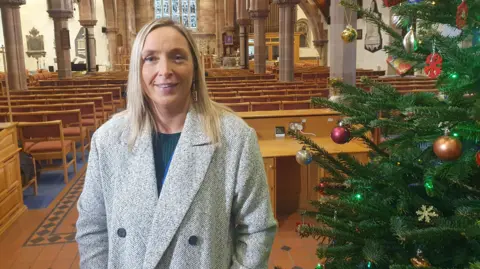 Mandy standing smiling at the camera. She is wearing a grey coat with black buttons. She has long blonde hair. There is a Christmas tree beside her.
