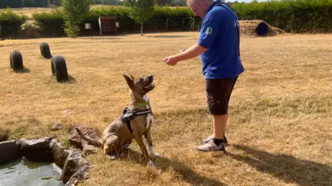 A photo of a man with a blue top and shorts standing up with a dog sitting on a floor next to pond.
