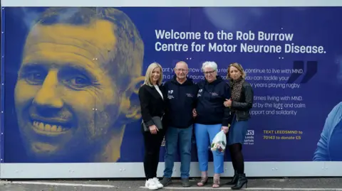 PA Media An older couple stand in front of a sign with a younger blonde woman either side of each of them. The older couple are both wearing navy hoodies with MNDA branding on. They are stood in front of a sign which reads 'Welcome to the Rob Burrow Centre for Motor Neurone Disease' and has a picture of Rob Burrow's face on it which shows him smiling.