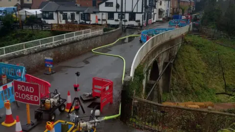 BBC A bridge with buildings on the opposite side. There are road closed signs and traffic cones and various pieces of machinery as well as blue temporary fencing on the bridge. There are fixed white rails either side of the bridge and a large grass embankment can be seen on the far side.