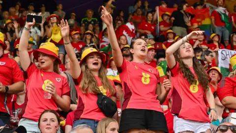 Four female Wales fans all wearing red shirts standing in the crowd and cheering at the match. Two of them are wearing red and yellow bucket hats. They are surrounded by other fans who are wearing red shirts. 