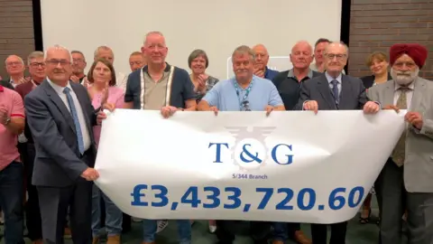 BBC A group of 16 Goodyear workers stand together holding a banner showing the figure £3,433,720.60