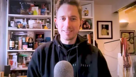 A man in a black hoodie sits behind a microphone in a room with a crowded shelf behind him