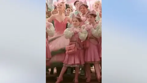 Universal Pictures Screen shot of Ariana Grande who is surrounded by a group of girls dressed in pink costumes. Lily is in the foreground.