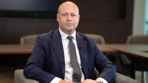 Dan Thomas sitting in a chair, wearing a blue suit, white shirt and pokadot tie.