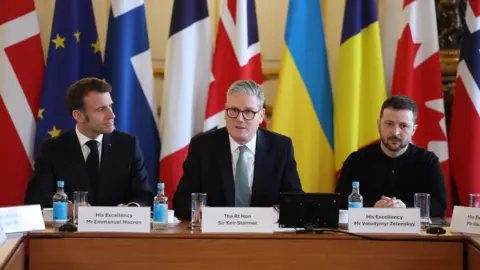 From left to right, Emmanuel Macron, Sir Keir Starmer and Volodymyr Zelensky sitting at a desk with the flags of the world behind them.