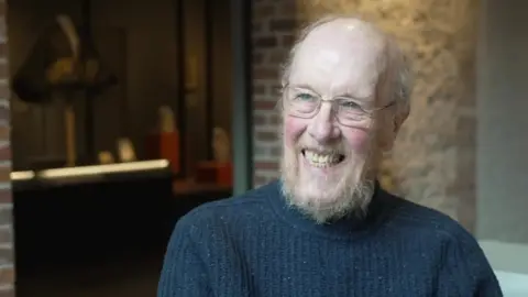 Peter Whittlesea / BBC An old man with a beard and glasses. He is sat in a bare brick-walled building.