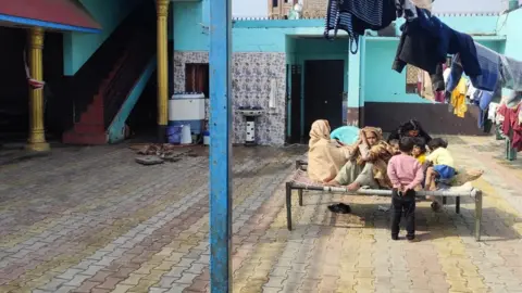 A quiet courtyard inside a village home in Umri. Several women and children sit together on a woven cot while one child stands nearby. Clothes hang on lines overhead, and the surrounding walls are painted blue and turquoise, with doorways and a staircase leading into the house
