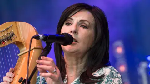 Moya Brennan singing into a microphone with a harp held in her right hand. 