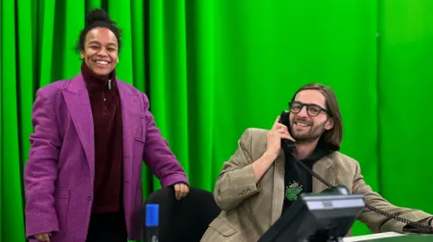 Jon Wright/BBC Mae Munuo is stood in a red zip-up fleece and purple coat. She is smiling at the camera and is stood in front of a green curtain. She is next to Jack Harrold. He is sat down at a desk holding a telephone to his ear and is wearing a tweed blazer. He is looking at the camera and smiling and has black-rimmed glasses on. 