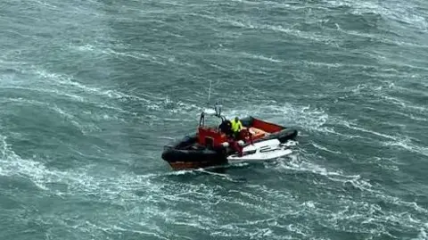 Lifeboat with white dinghy close to its side in choppy seas.