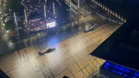 MICH2255 Two helicopters are hovering above the piazza at Media City, with the corner of a building seen nearby and a giant video screen and a tram stop also nearby.