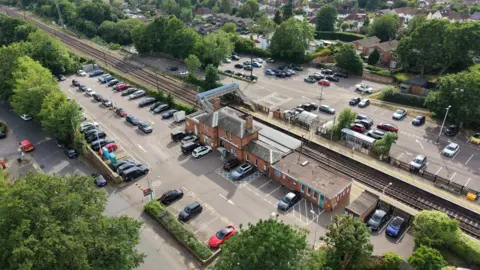 Ant Saddington/BBC An aerial view of a railway station and surrounding car parks. A red‑brick station building sits near train tracks, with platforms on either side connected by a covered footbridge.