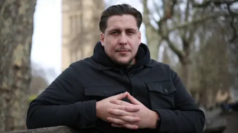 Reuters A man in a black coat. He is sat on a bench on a grey day. 
