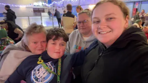 Family photo A family photo with Oliver at the centre with his grandma, mum and sister. There is an ice rink behind them.