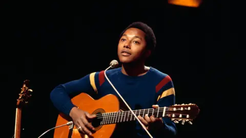 Getty Images A man with short dark curly hair, a blue jumper and a wooden acoustic guitar, sitting in front of a microphone with a black background