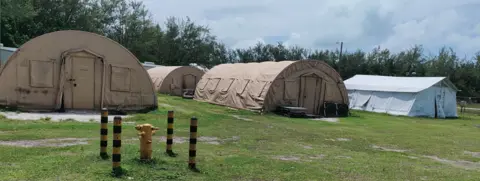 Photo shows 3  circular  beige tents and a achromatic  thin   connected  greenish  grass. In the inheritance  determination   are trees