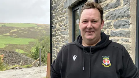 Aled Wyn Davies wearing a black hoodie and standing in front of a house with green hills in the background