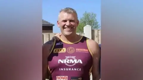 A man with short grey hair looks directly at the camera, wearing a burgundy running vest with yellow trim. The vest has sponsorship logos on the front. It is a sunny day and there is a wooden panelled fence and a tree behind him.