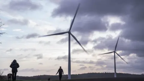 Two tall, white wind turbines are spinning in the wind. They are in a field with a line of trees in the distance. Two people in the foreground are walking their dogs.
