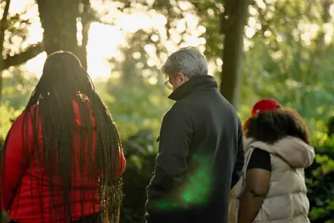 Fergal walks betwixt  2  South African asylum seekers. Seen from behind, the pistillate   connected  the near  wears a reddish  coat, she has agelong  braids - the pistillate   connected  the close    wears a achromatic  sleeveless puffa overgarment   and reddish  shot   headdress  implicit    agelong  escaped   hair