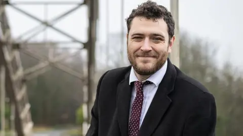 Hexham MP Joe Morris, wearing a black overcoat, shirt and tie and smiling with an out of focus bridge behind him.