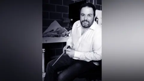 Pierre Bohanna Black‑and‑white photo of Pierre Bohanna seated on a stool in a workshop, holding a handcrafted wand, created for the Harry Potter films.