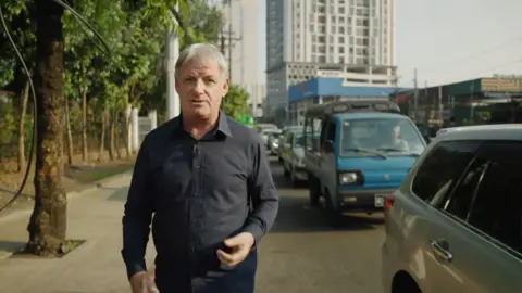 BBC correspondent walking next to queue of vehicles in Myanmar