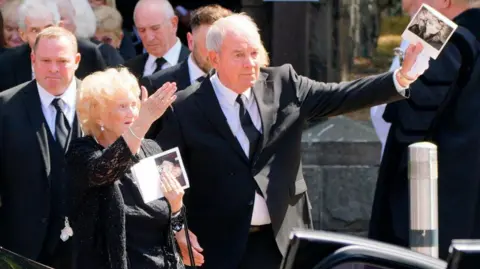 PA Media A man with short grey hair and wearing a white shirt, black tie and black suit is looking in front of him and waving as he comes out of a church. An older woman with blonde hair and a black outfit is also waving.