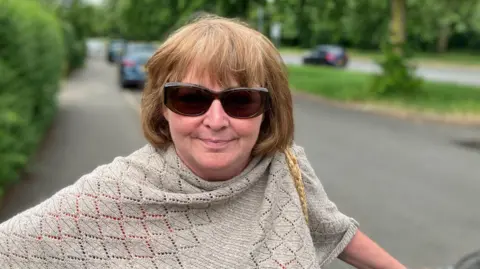Sam Read/BBC Elaine Jones looks directly at the camera and smiles. She is standing on a pavement and holding the handlebar of a bicycle in her left hand. She has auburn/grey hair and dark brown sunglasses. She is wearing a beige/grey shawl.