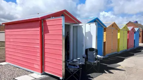 East Riding of Yorkshire Council A row of brightly painted beach huts in red, blue, orange and yellow. One has its front patio doors open and two camping chairs outside. They sit on concrete slabs and gravel.