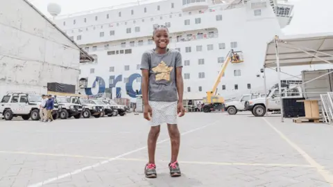 Mercy Ships Fudia, after her surgery, she stands outside in a different section of the dockyard, with a white and blue Mercy Ship in the background, as well as a number of parked white jeeps. Fudia wears her hair in braids, with a slate grey short sleeved cotton t shirt and a gold foil star on the front, she has pale pink capri shorts and wears black platform sliders. She is smiling, with her legs straighter and now wears black trainers with neon pink laces.