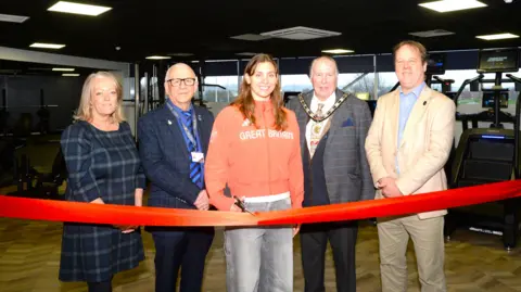 South Kesteven District Council Five people - two women and three men - stand behind a red ribbon in a gym surrounded by treadmills and weights. A woman in the centre of the image wears a red tracksuit top featuring the words "Great Britain". She is holding a pair of scissors as the other people dressed in suits smile into the camera.
