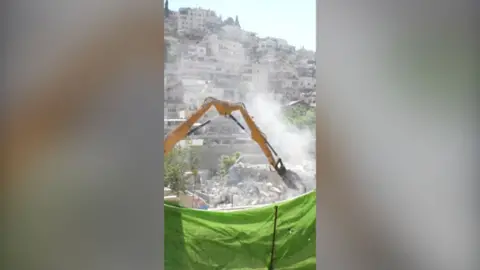 A yellow bulldozer engaging in the demolition of homes. It is behind a raised green net and behind it are other houses.