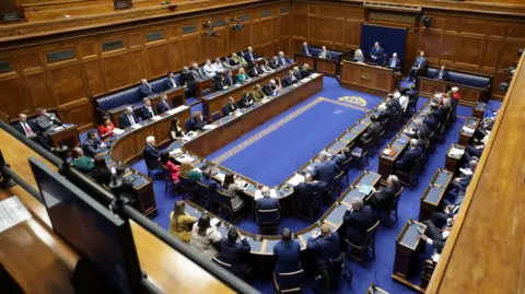 The inside of the Stormont Assembly chamber. There are three benches with people sitting on them. The walls are wooden panelling and the carpet is blue.