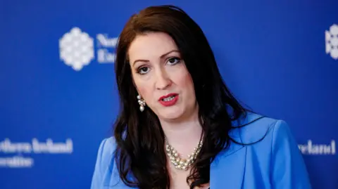 Emma Little-Pengelly is mid-speech as she is photographed. She is standing in front of a blue background with the Northern Ireland Executive logo on it. She has long, dark hair and she is wearing a blue suit jacket, a pearl necklace and pearl earrings.