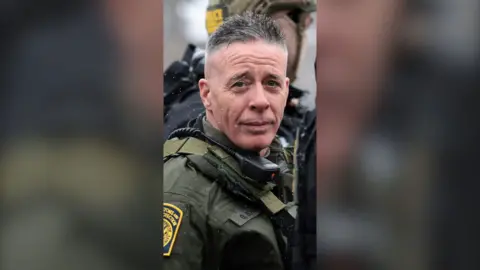 A man, border patrol commander Greg Bovino, in uniform looking at the camera.