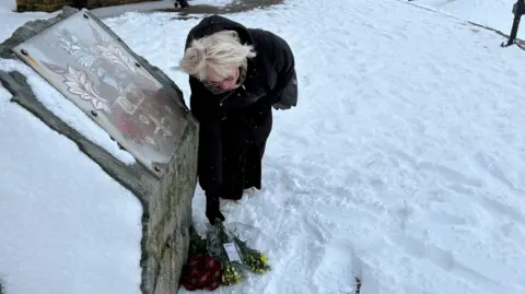 Kevin Shoesmith/BBC A grey-haired woman, wearing spectacles and dressed in a long coat, lays a simple bouquet of flowers next to a monument in front of the pier towers, a Withernsea landmark, on the seafront.