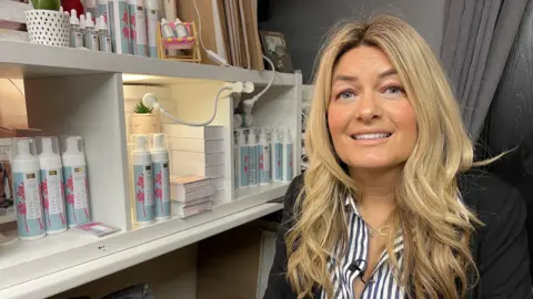 Shoned Owen, with blonde hair, a striped shirt and a black blazer, sits on the right of frame smiling at the camera. To the left are bottles of her tanning products displayed on white shelves.