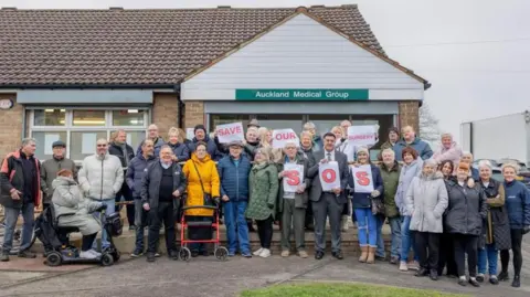 About 30 people are standing outside St Helens surgery owned by the Auckland Medical Group. Some of them are holding pieces of paper which say Save our Surgery, another three are holding the letters SOS up.