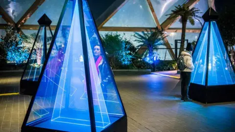 Dreamy Place CGI images of three blue isosceles pyramids. They are made of glass and filled with blue lights. There are in an empty room. Two people are looking into two of them. There are triangular windows on the back wall which show the sky through them. The sky is blue 