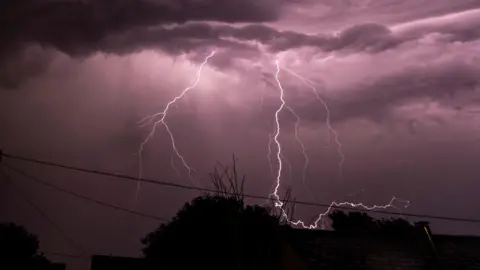 A lightning bolt pictured over Winterslow in Wiltshire. The sky is a purple tone from the flash. The silhouettes of trees and power cables can be seen below.