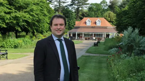 Bedford Lib Dems A man standing in a park with a pavilion behind him. He is wearing a black suit with a green tie and a light blue shirt.