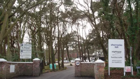 Geograph/Norman Caesar A brick wall entrance with iron gates opens to the former nightclub site. The sign reads Shorrocks Hill nightspot Please respect our neighbours. The area is surrounded by woodland and tall trees. 