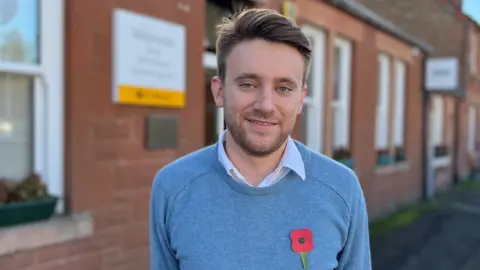 A smiling man with reddish brown hair and a beard wearing a blue jumper and a poppy stands in front of a sandstone property