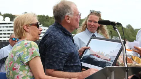 Several people stand at an outdoor podium during a ceremonial event by the waterfront. Alan speaks into a microphone while holding a framed photograph of a red lifeboat at sea. Annette stands beside the podium, and additional RNLI crew members are visible in the background. A large vessel and trees can be seen behind the group under an overcast sky.