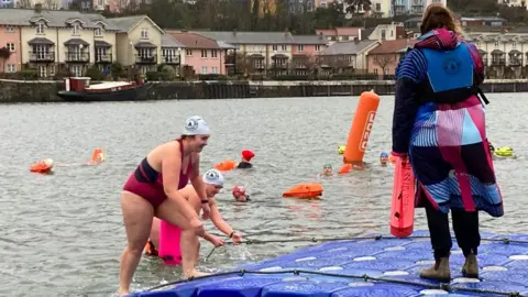 Two swimmers using grab ropes to leave the water, while a lifeguard watches on